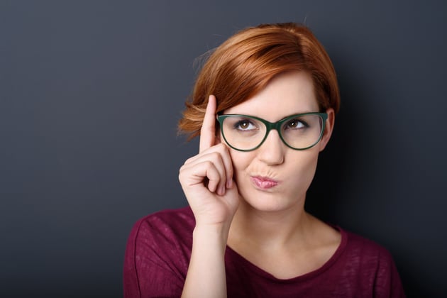 Nerdy scholastic young woman wearing geeky glasses standing thinking with her finger raised and a grimace of concentration in a humorous stereotypical depiction, over a dark background with copyspace-1